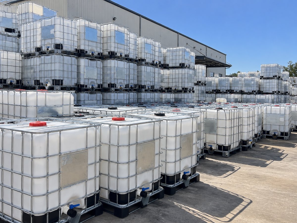 Graded IBC totes lined up in the yard outside the plant, clean white bottles in galvanised cages on black plastic pallets.
